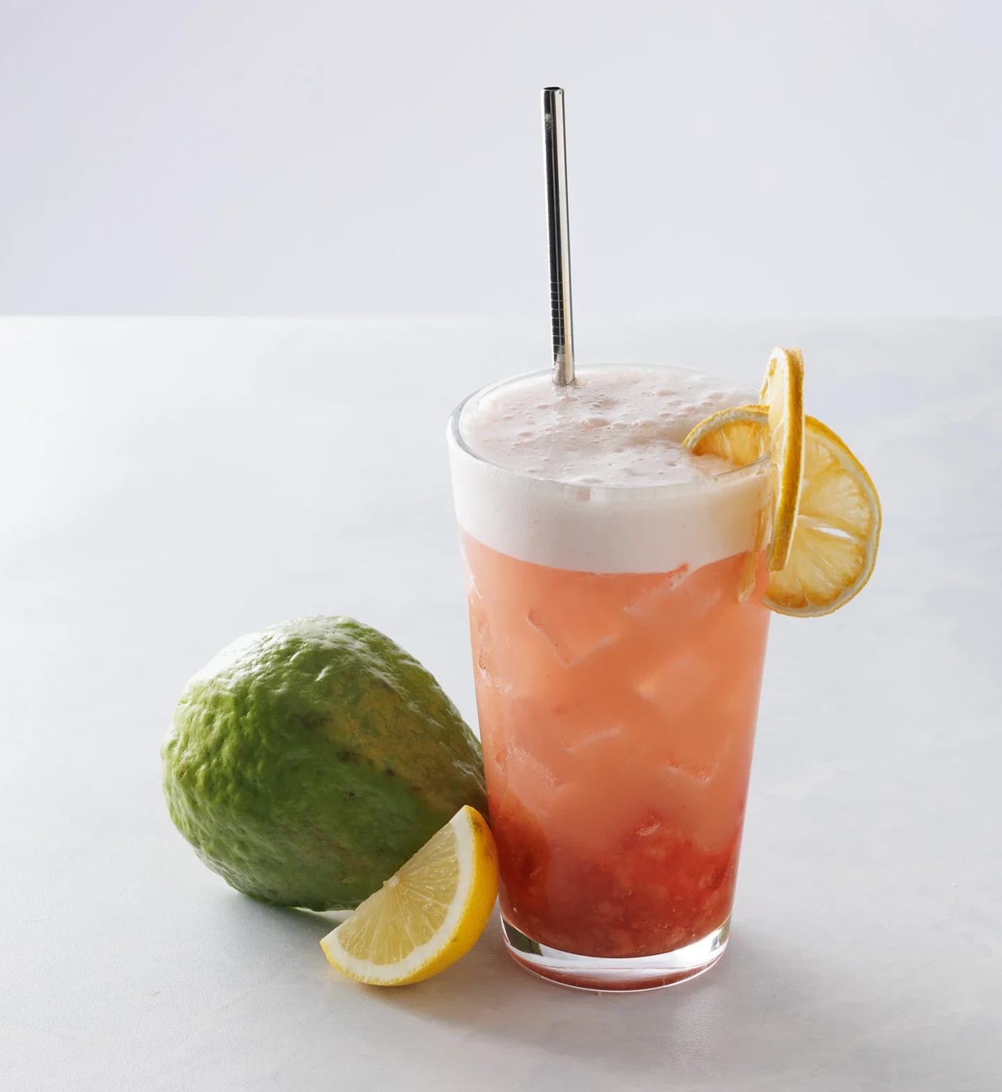 Pink guava lemonade in a glass with gauva and lemons next to it.